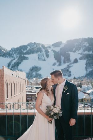 Romantic winter wedding portrait with snowy mountains and brick building backdrop.