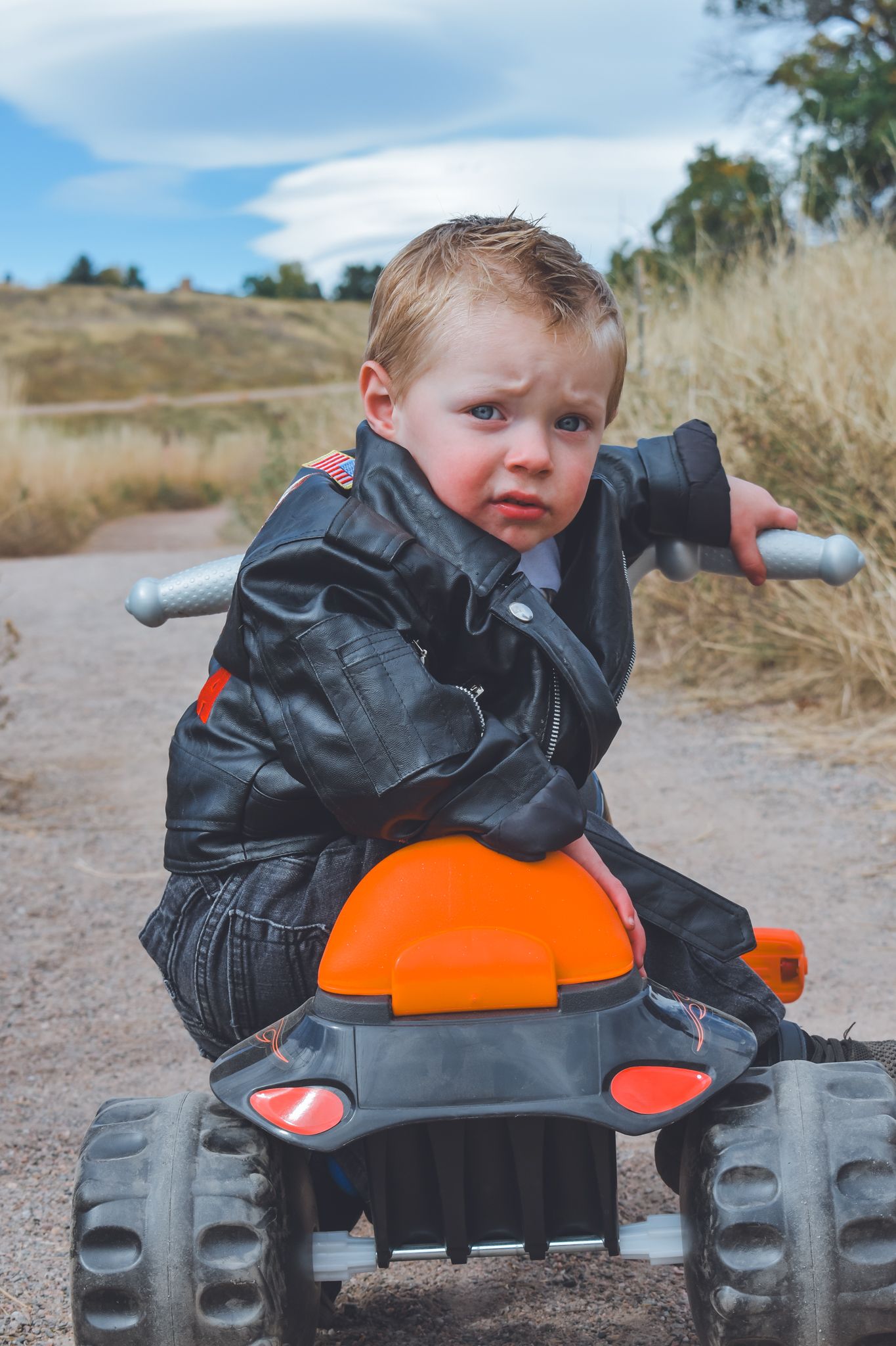 Toddler boy in a leather jacket sitting on an orange and black toy ATV on a dirt path outdoors.