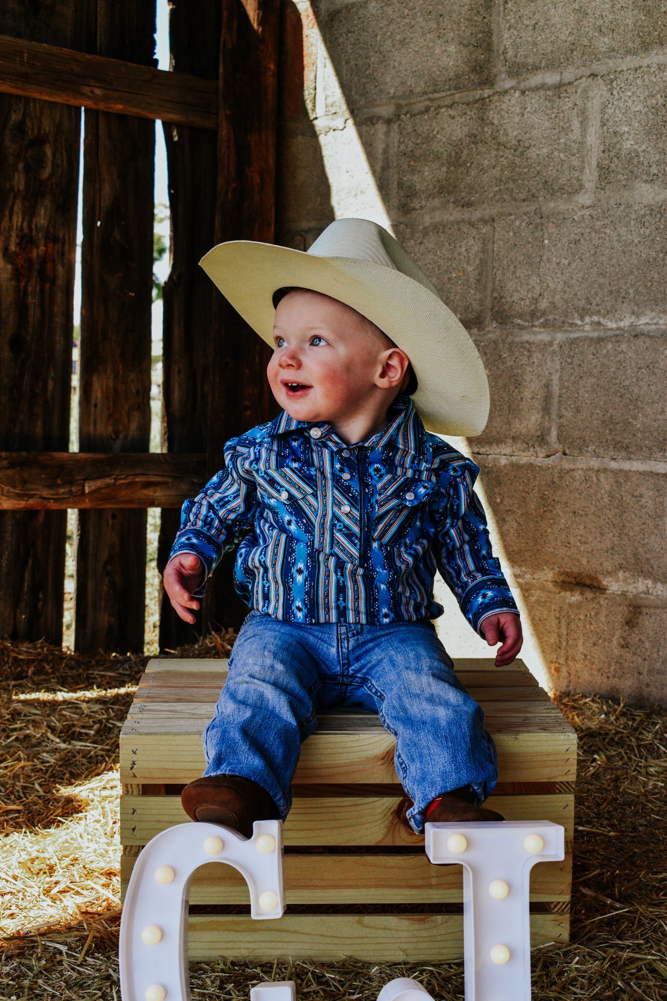 Smiling toddler boy wearing a cowboy hat and blue western shirt sitting on a wooden crate in a rustic barn.