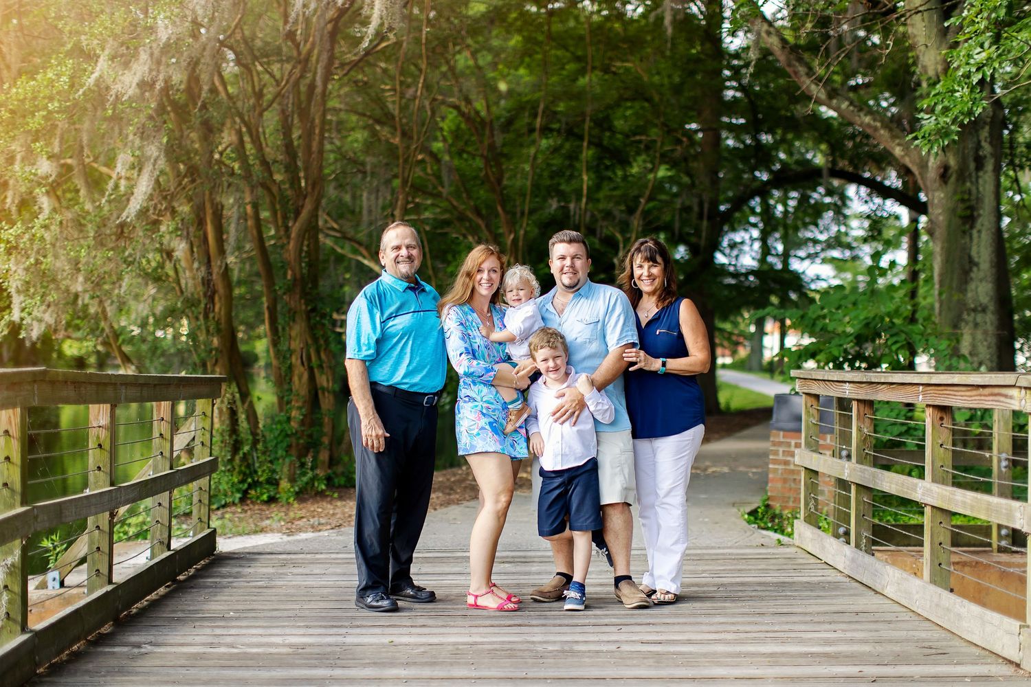 A family poses together on a wooden bridge surrounded by lush green trees on a sunny day.