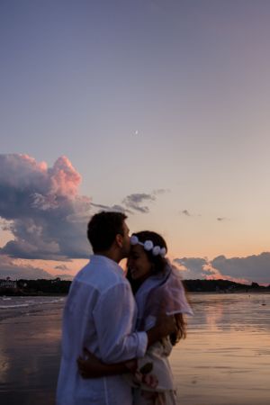 A man kisses a woman's forehead while standing on a beach at the water's edge as the sun sets and clouds fill the sky.