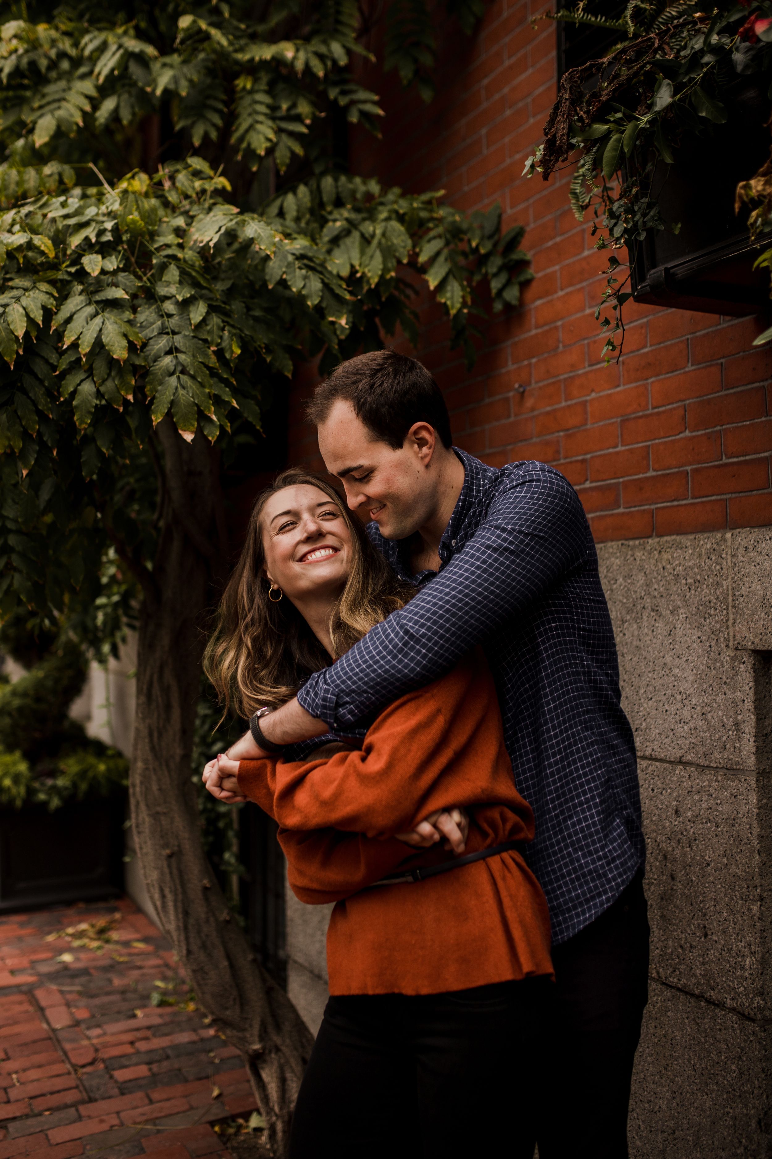 A man wraps his arms around a woman, both facing the camera, as they rock back and forth in front of a brick wall.