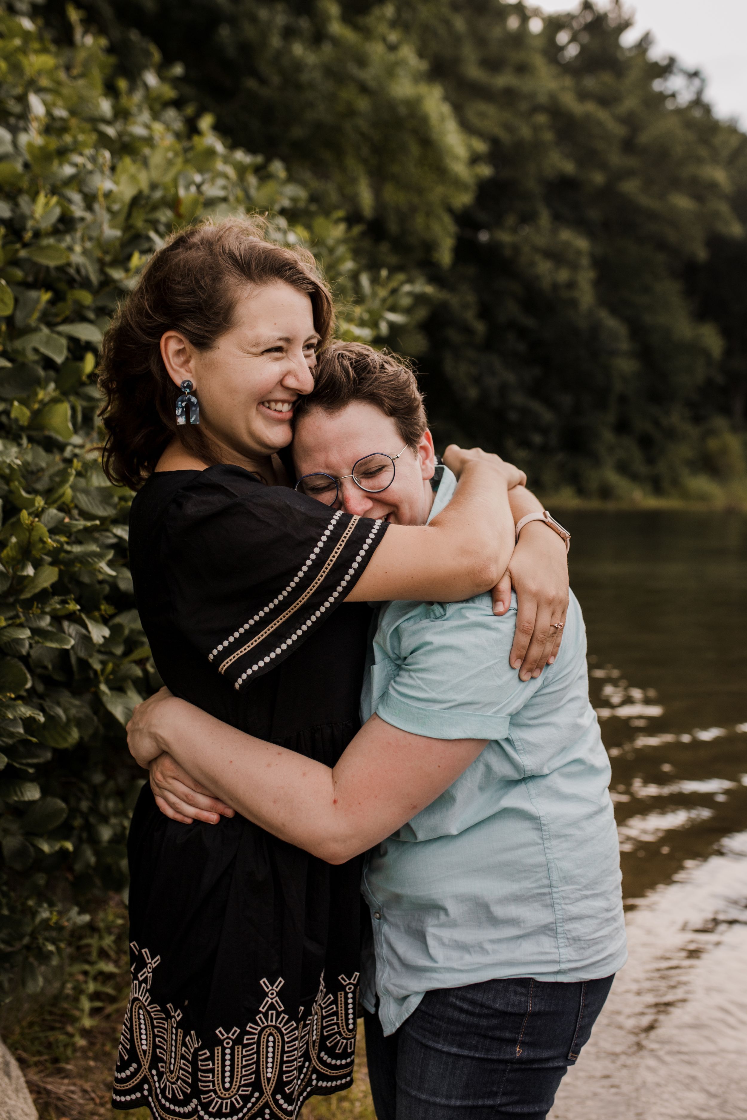 A couple tightly embraces while smiling and surrounded by trees and a lake..