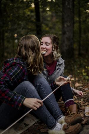 Two women sit on the ground in front of a campfire and take a bite of the same s'more.