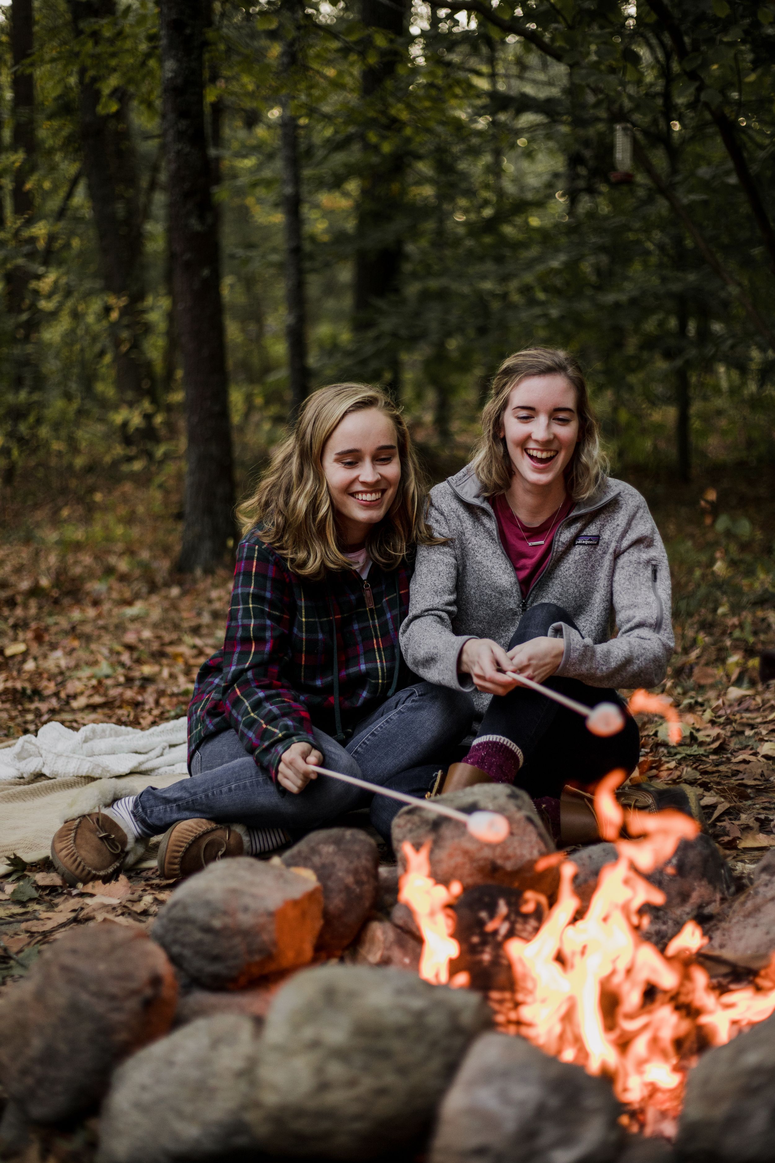 A couple sits in front of a fire while roasting marshmallows on sticks in the woods.
