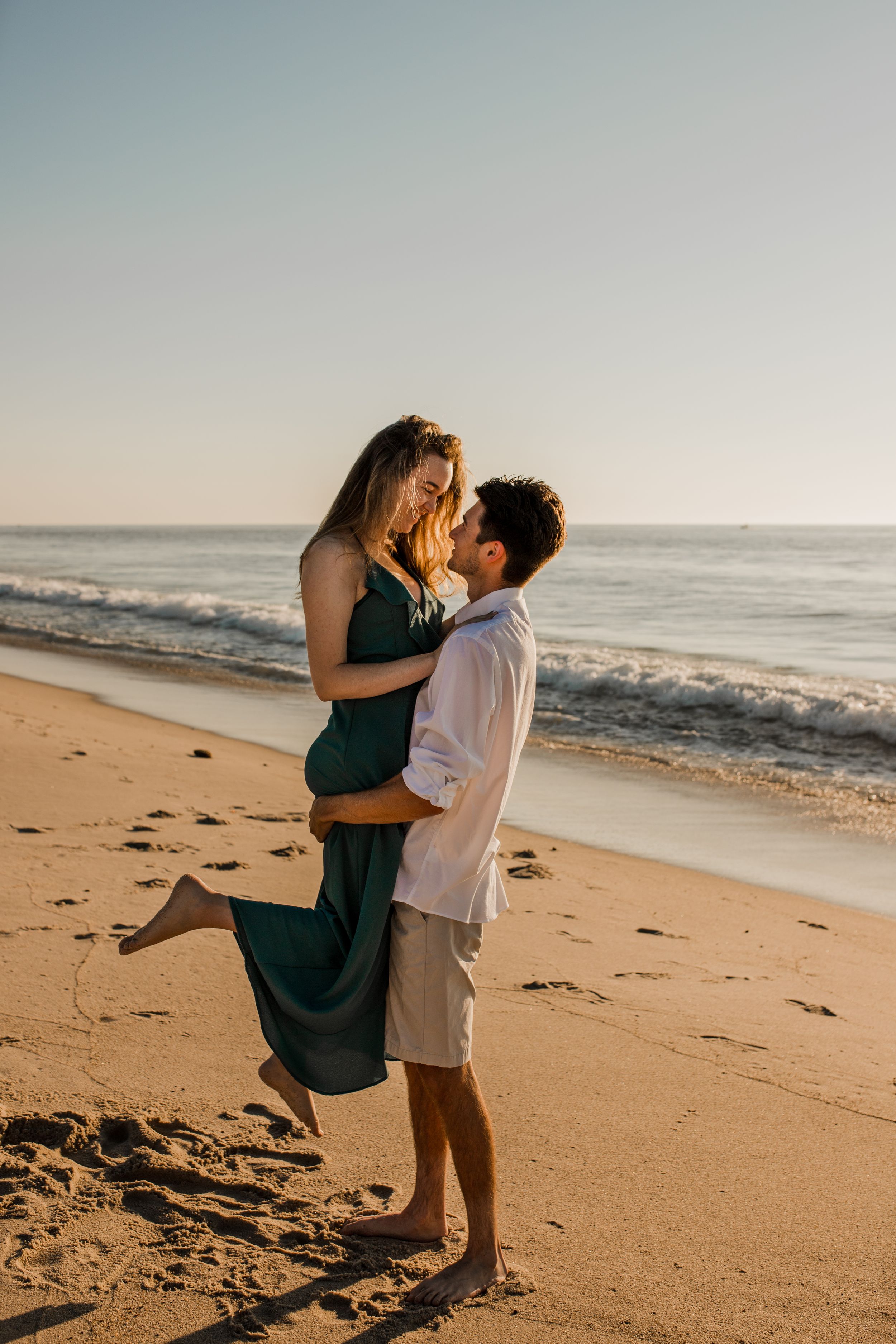 A man picks up a woman on a beach as they stare into each others' eyes and the woman pops her leg.