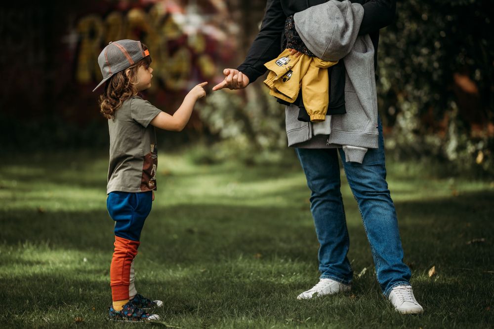 little boy wearing a baseball cap backwards, arm out touching fingers with a man in front of him