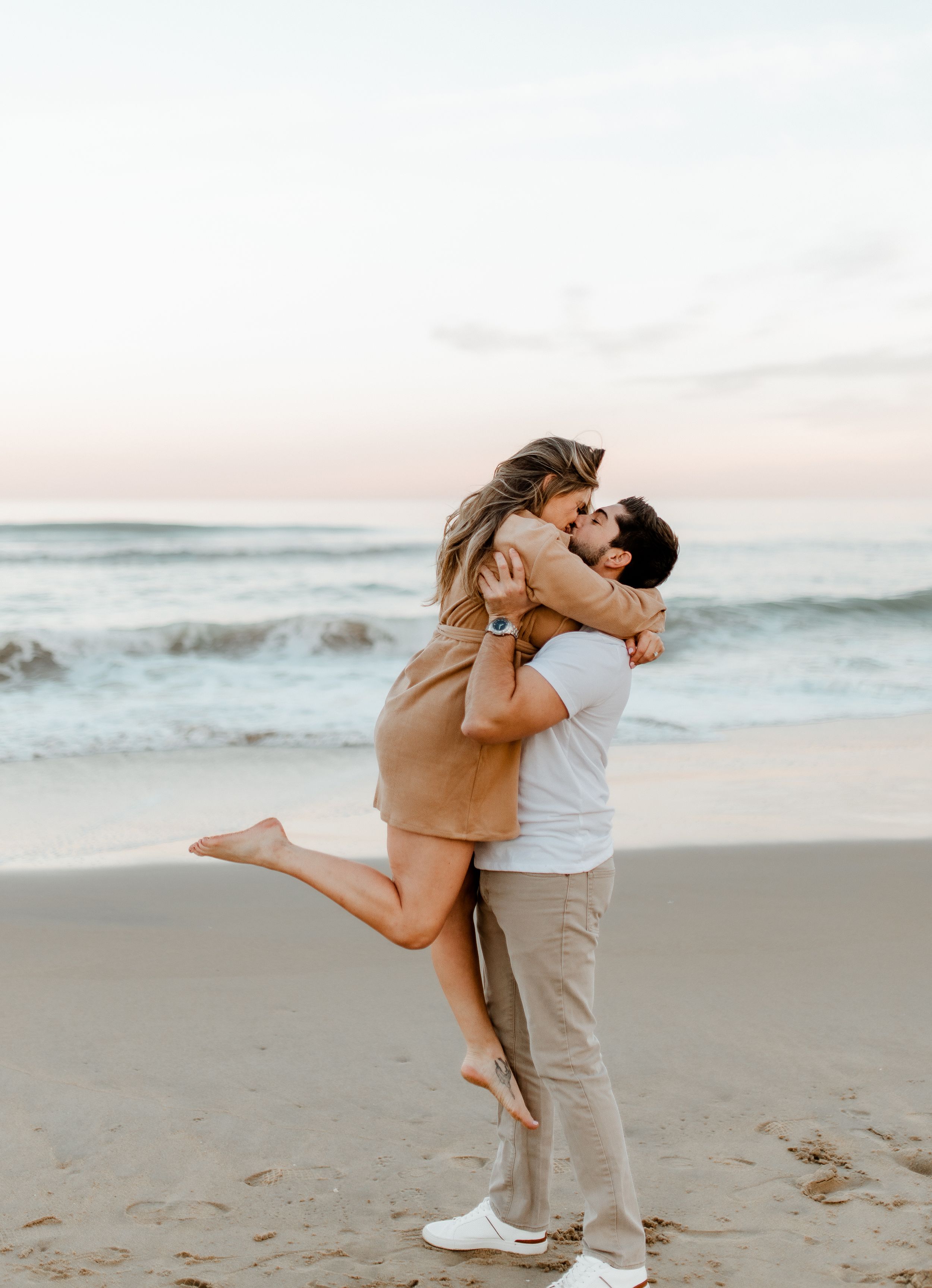 Romantic couple embracing on beach at sunset with waves crashing in background.