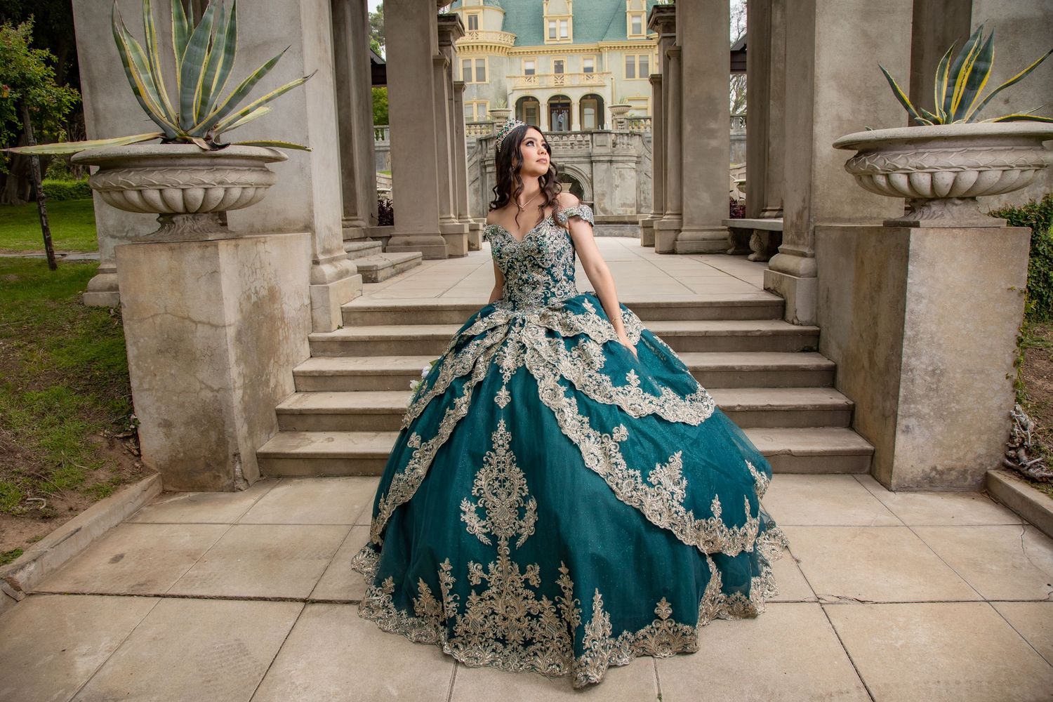 Young woman in a teal and gold quinceañera ball gown posing on stone steps of a grand building.