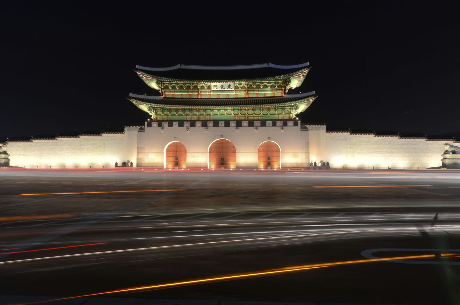 Illuminated Gyeongbokgung Palace gate glows at night with light trails from passing traffic.