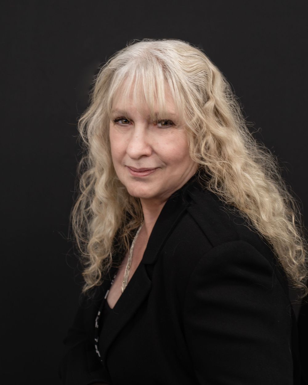 Professional headshot wearing black blazer against dark background with wavy blonde hair.