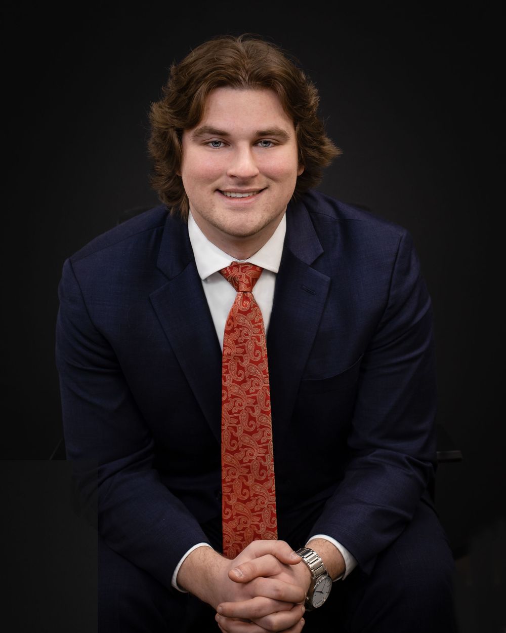 Studio portrait in navy suit with patterned red tie against dark background.