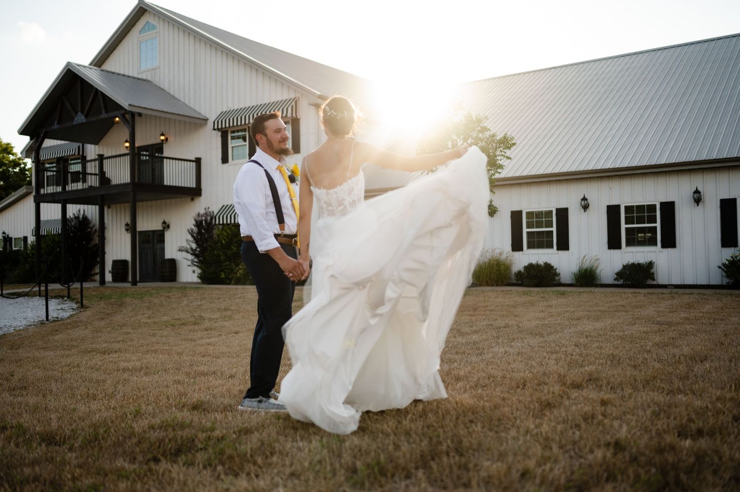 Wedding couple silhouetted against a white barn at golden hour as bride kicks up her foot playfully.