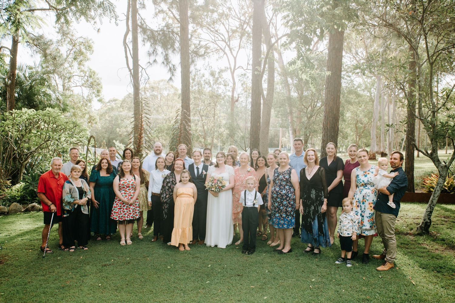 A large group wedding photo taken in a sunlit garden with tall trees in the background.