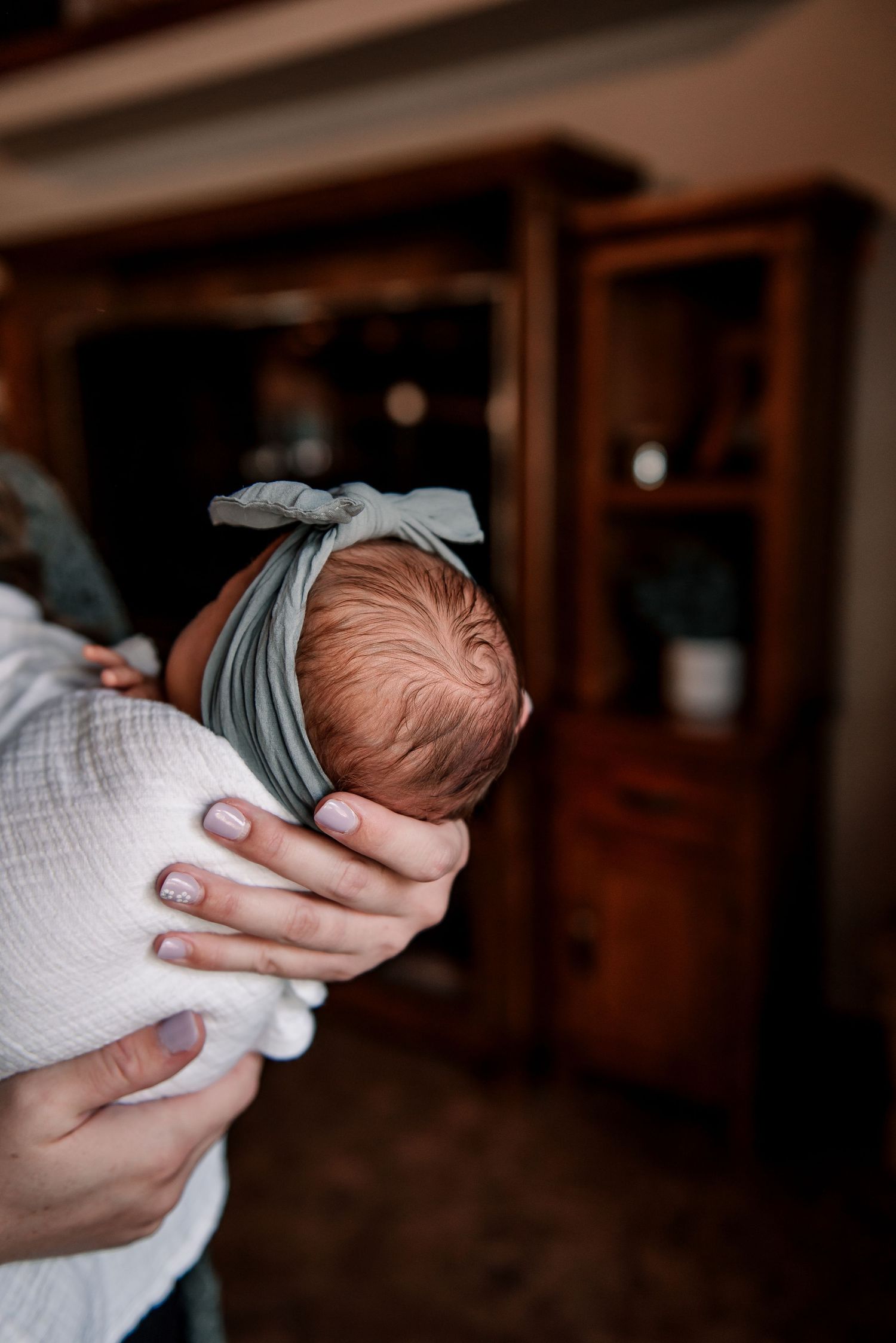 Newborn baby wearing a grey bow headband being cradled in white swaddle blanket.