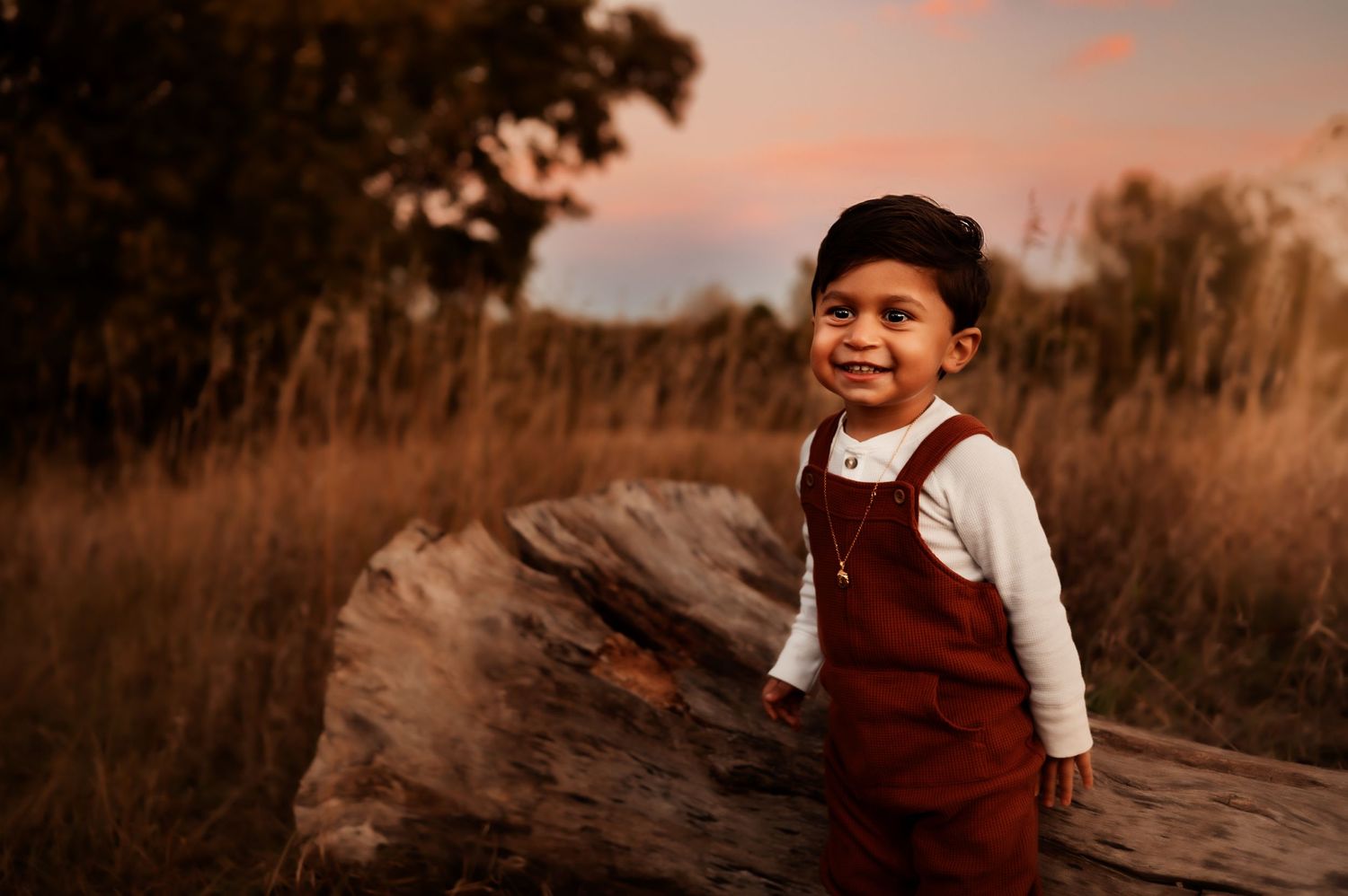 Child in red overalls smiles during outdoor sunset portrait session.