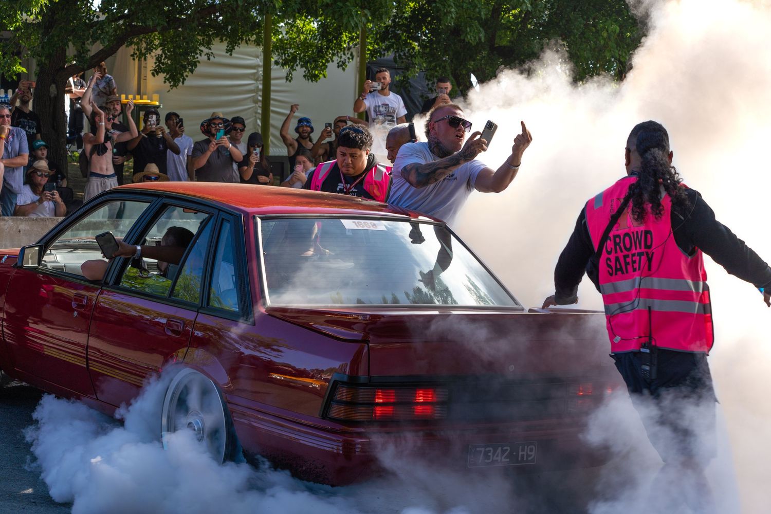 Red car performing a burnout with smoke and spectators at a car event.