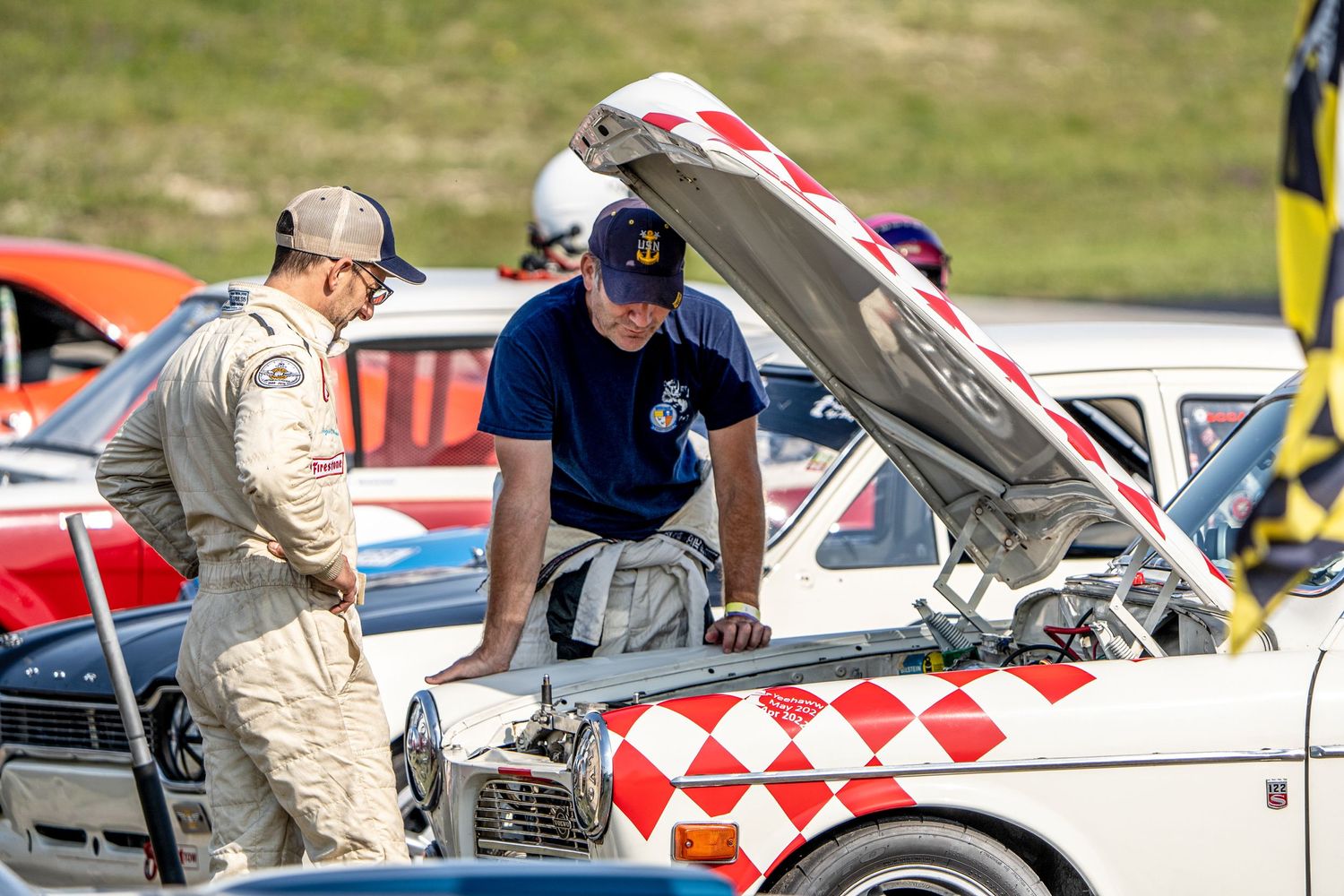 Two mechanics inspect under the hood of a vintage race car with checkered pattern paint scheme.