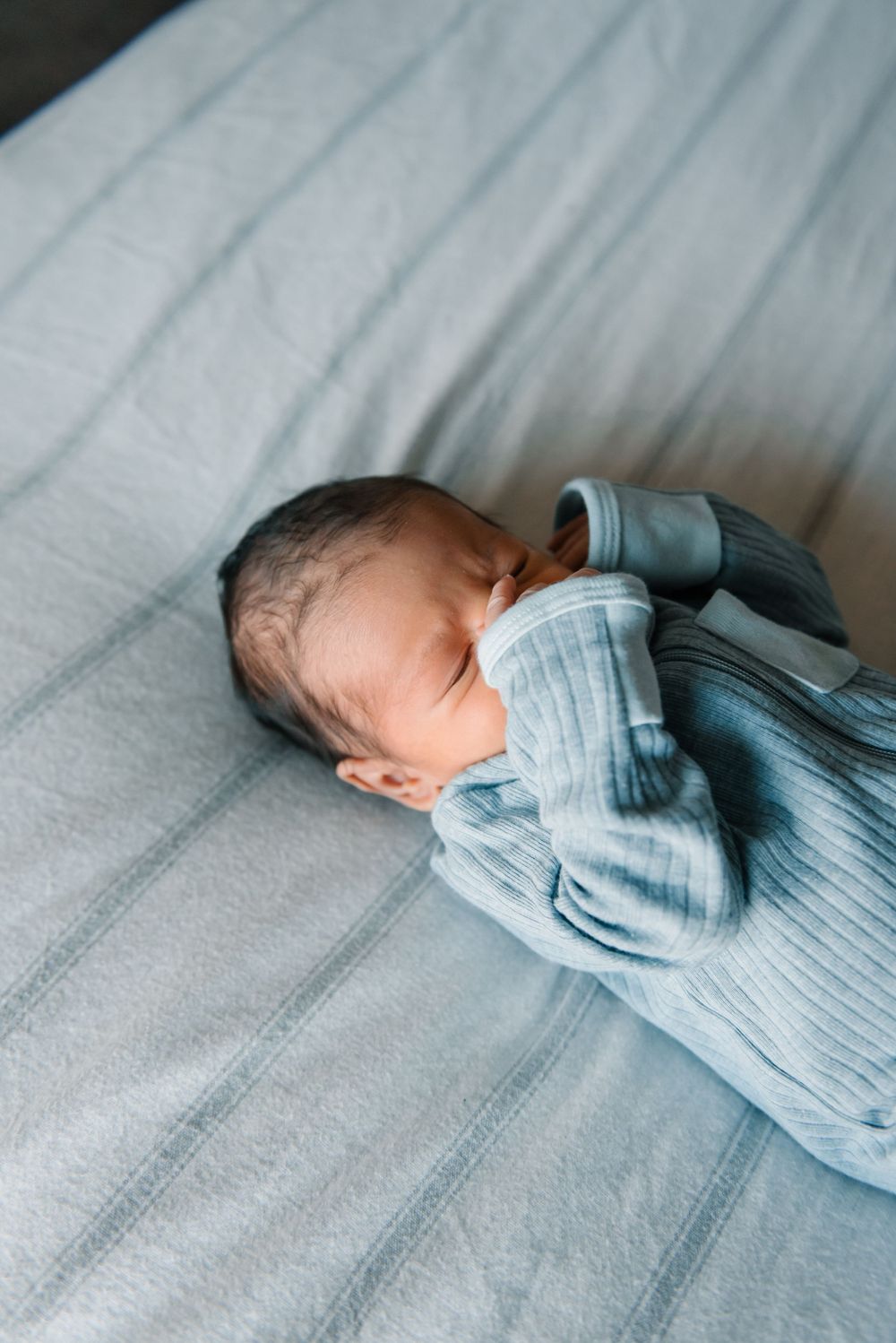 Sleeping newborn baby in light blue clothing resting on grey bedding.