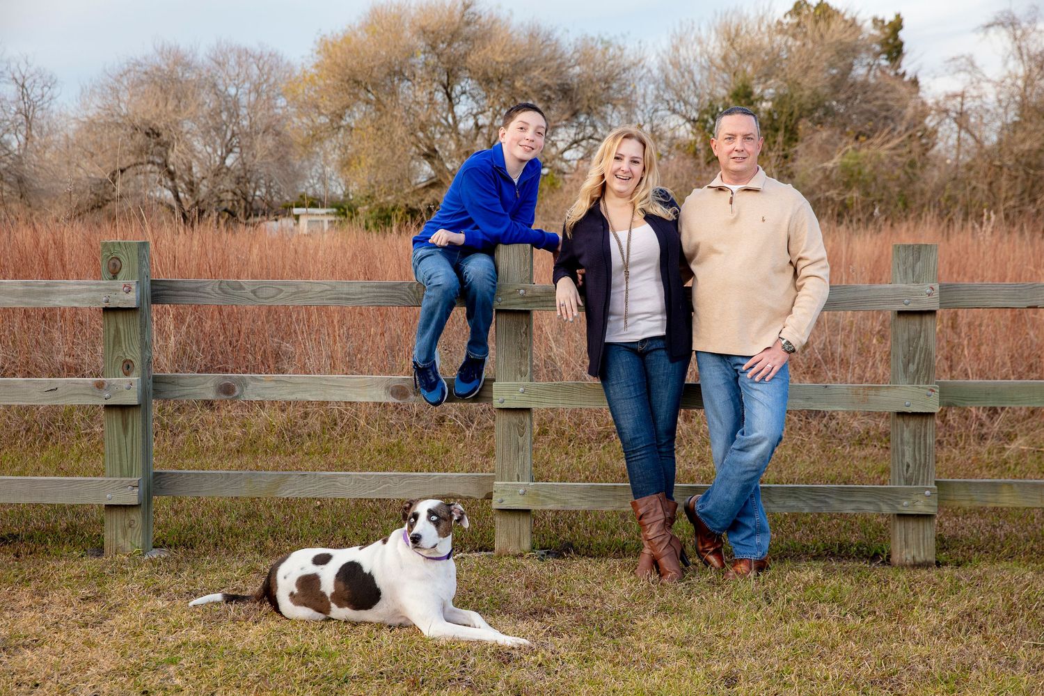 Family poses on wooden fence with spotted dog lying below.