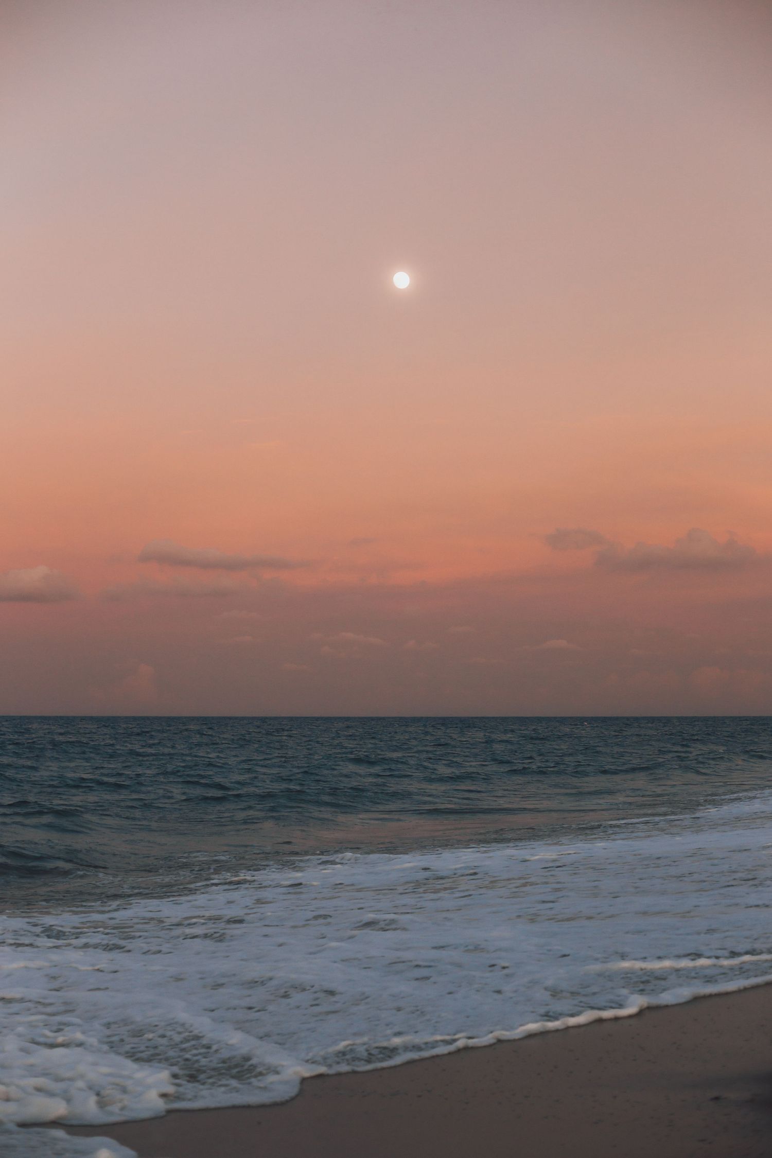 Peaceful sunset over the ocean with a glowing moon in the pink and orange sky.