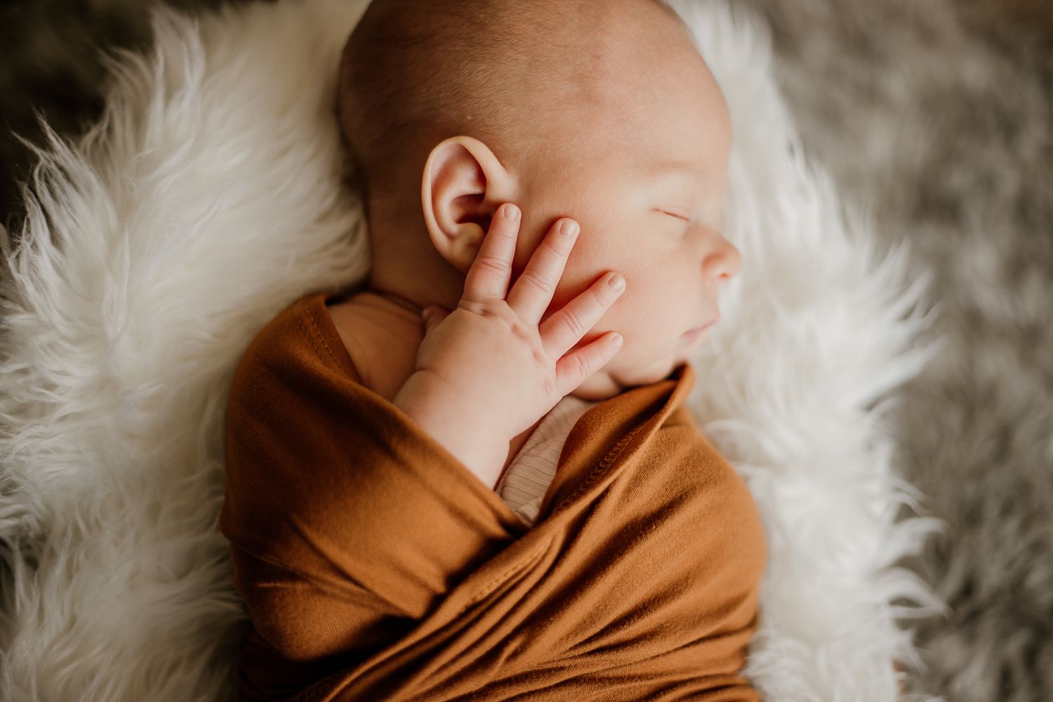 Artistic newborn photography with baby wrapped in brown fabric on white fur backdrop.