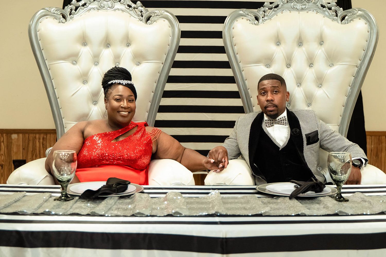 Couple sitting together on ornate white throne chairs against a striped backdrop.