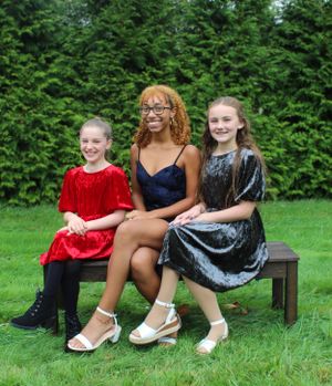 Three friends in formal dresses pose together on wooden bench with evergreen hedge backdrop.