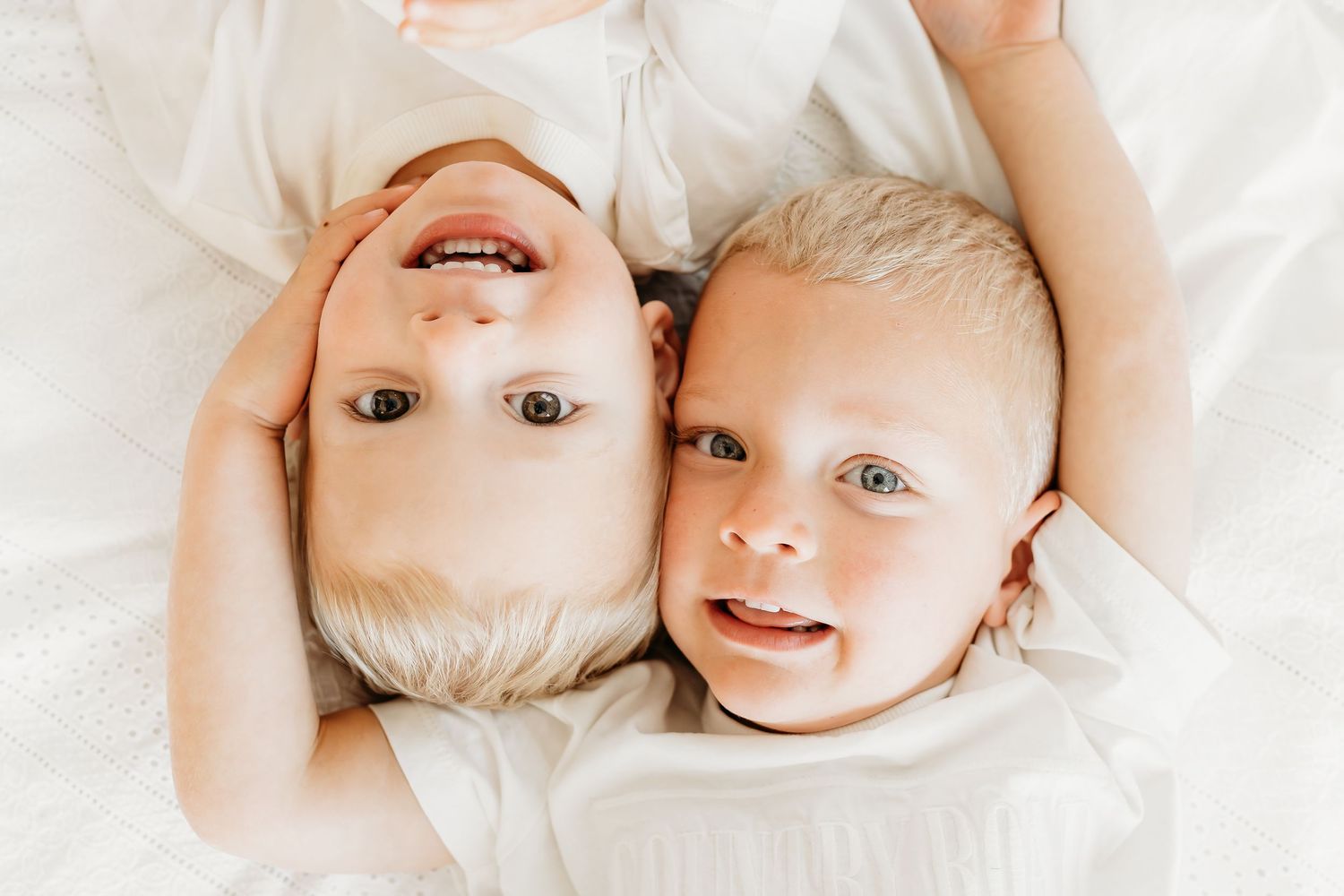 Two young children lying on white bedding together with joyful expressions.