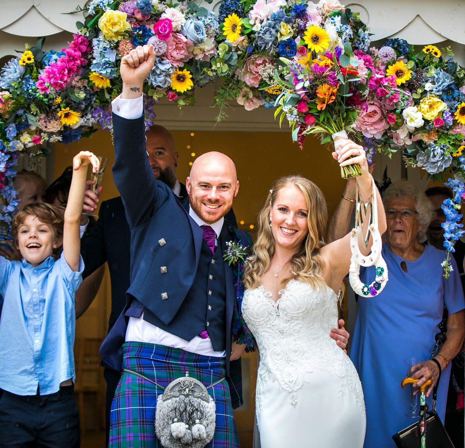 Wedding party celebrating under a floral arch with bright yellow and pink flowers.