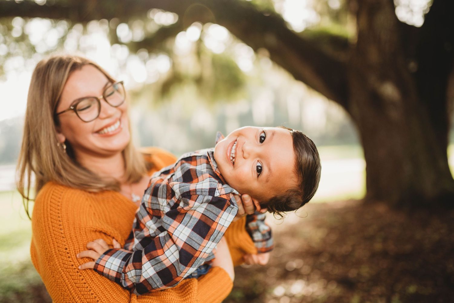Parent and child share joyful moment under tree with warm sunlight filtering through.