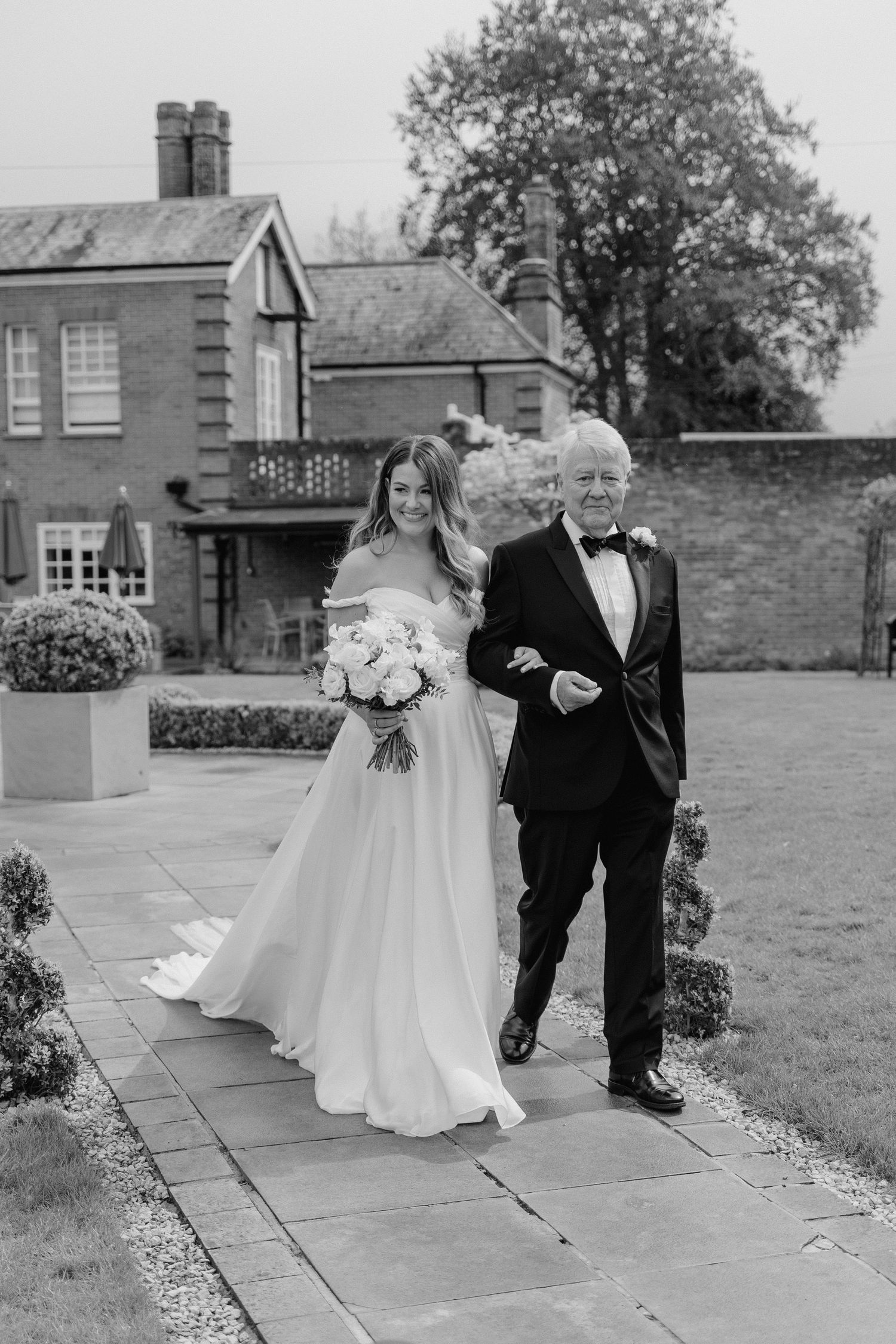 A black and white image of a bride being given away by her dad. they are walking through a garden.