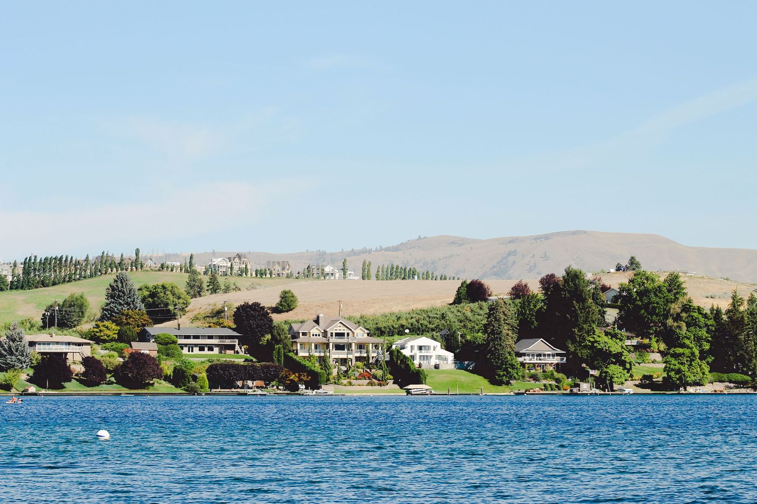 Lakefront homes nestled among trees with mountains in the background on a sunny day.