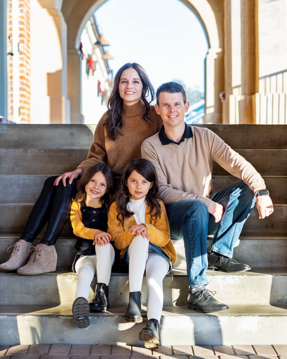 Orange County photographer captures Family of four with two young girls sitting on outdoor steps near decorated archways.