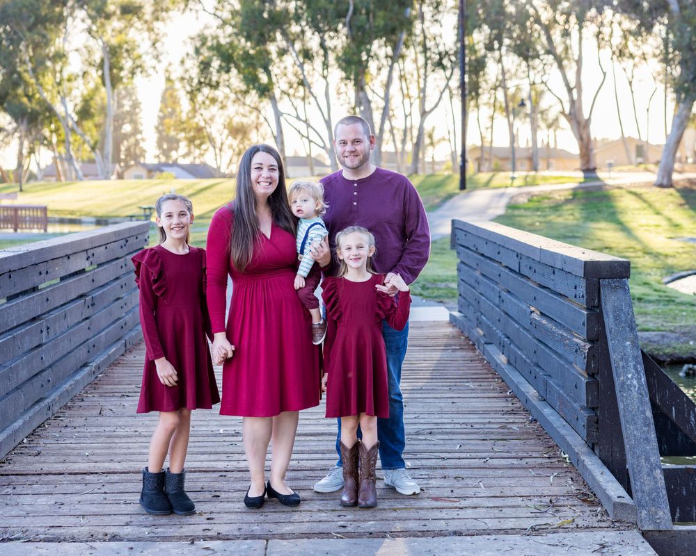 Orange County photographer captures Family of five in matching burgundy outfits posing on a wooden bridge in a park during golden hour.