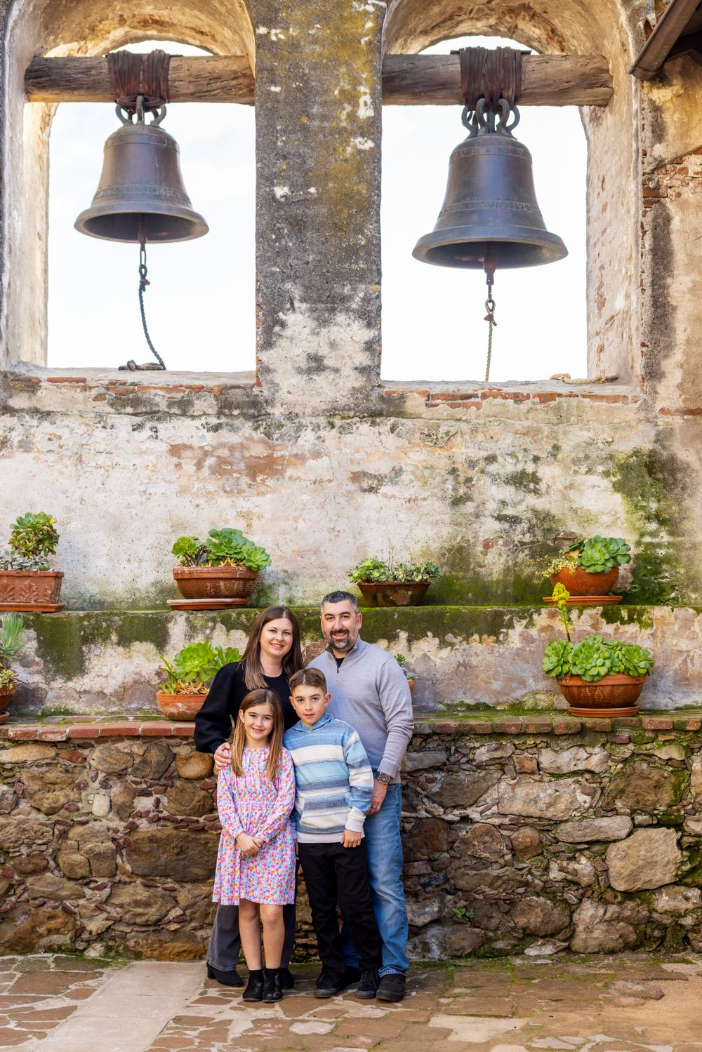 Orange County photographer captures Family of four posing under historic mission bells with stone walls and potted plants backdrop.