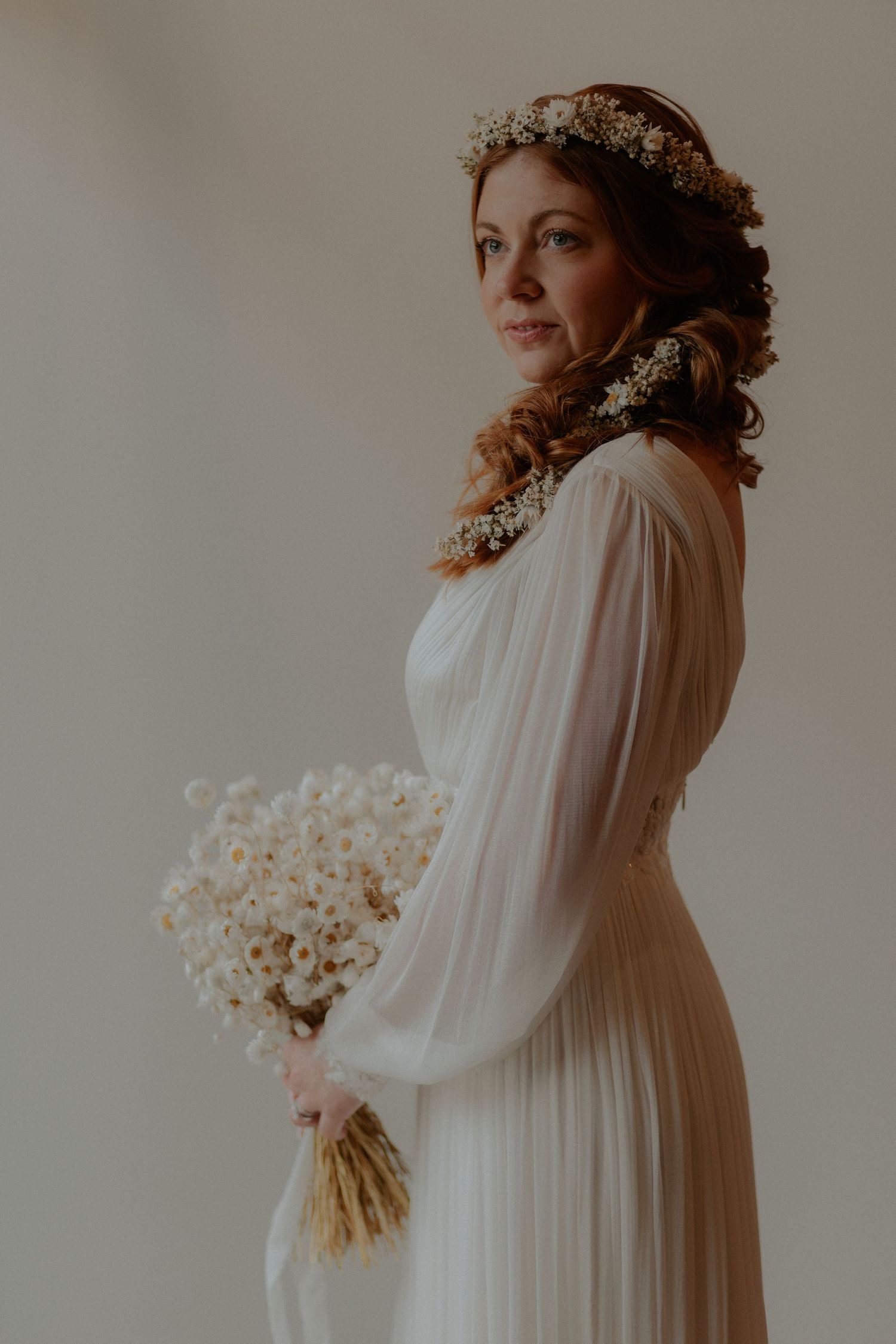 A bride in a flowing white dress with long sleeves and floral crown poses elegantly against a neutral backdrop.