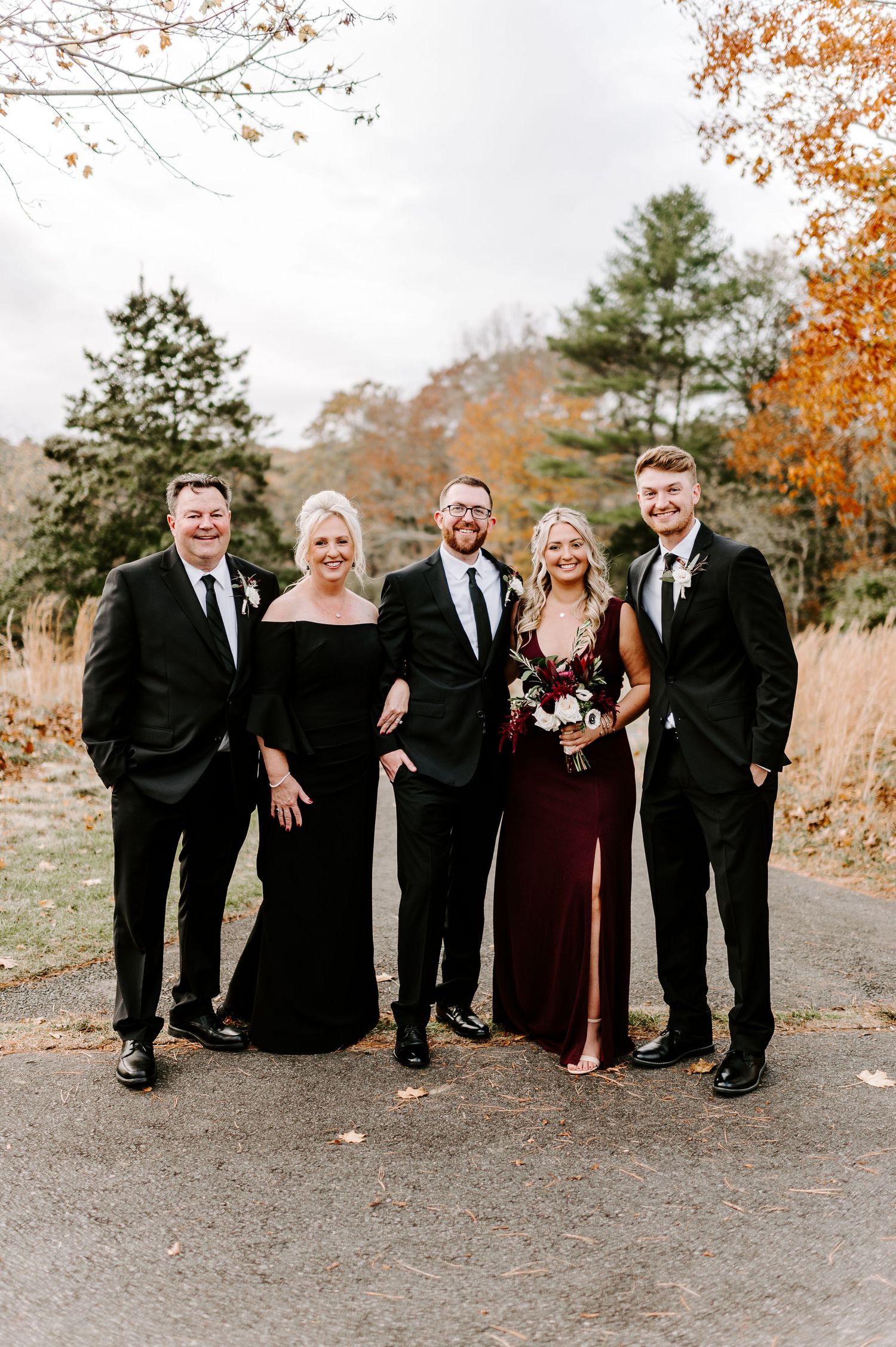 Wedding party members in black and burgundy formal wear pose together during fall celebration.