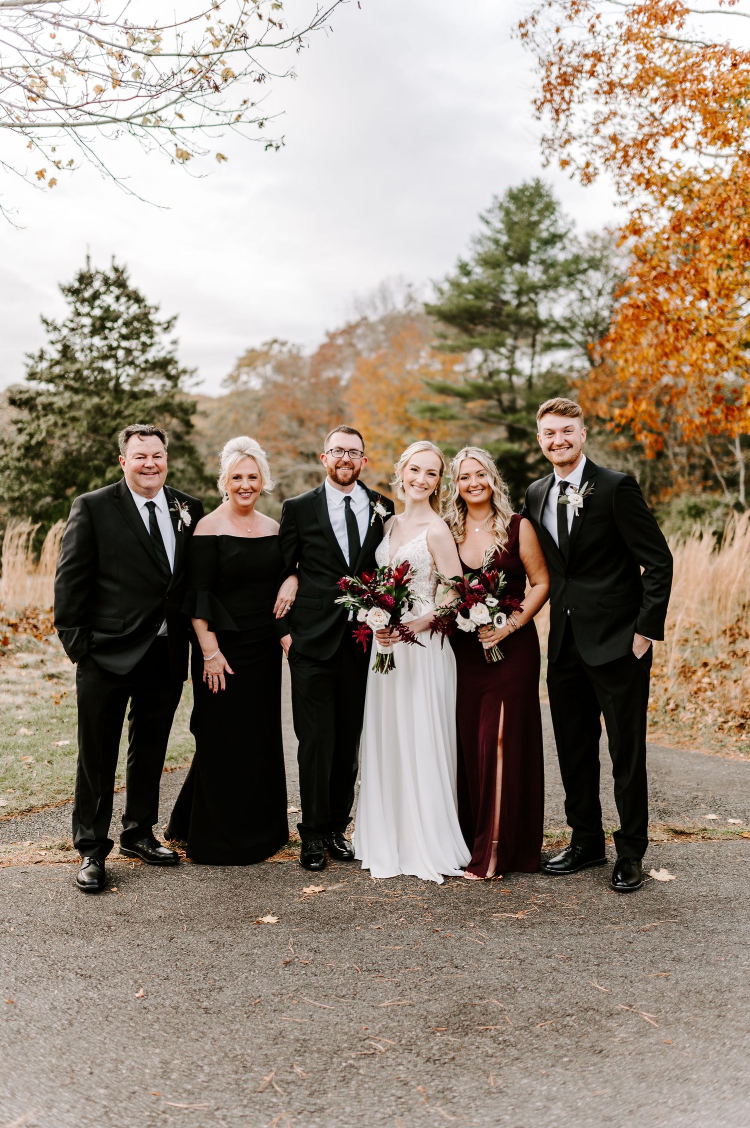 Wedding party in formal attire poses together on path with autumn scenery.
