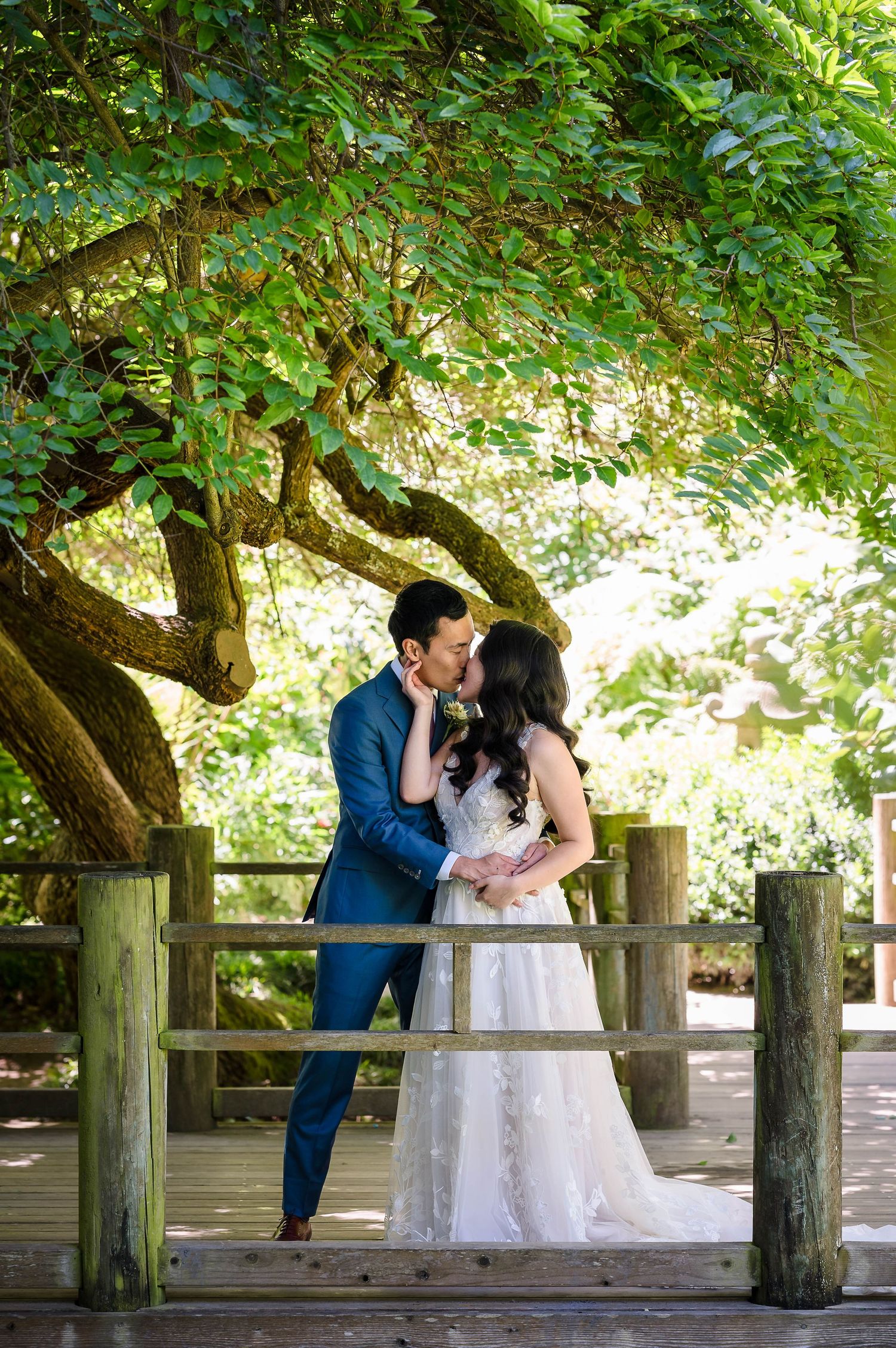 A tender moment shared between newlyweds on a wooden bridge under a canopy of green leaves.