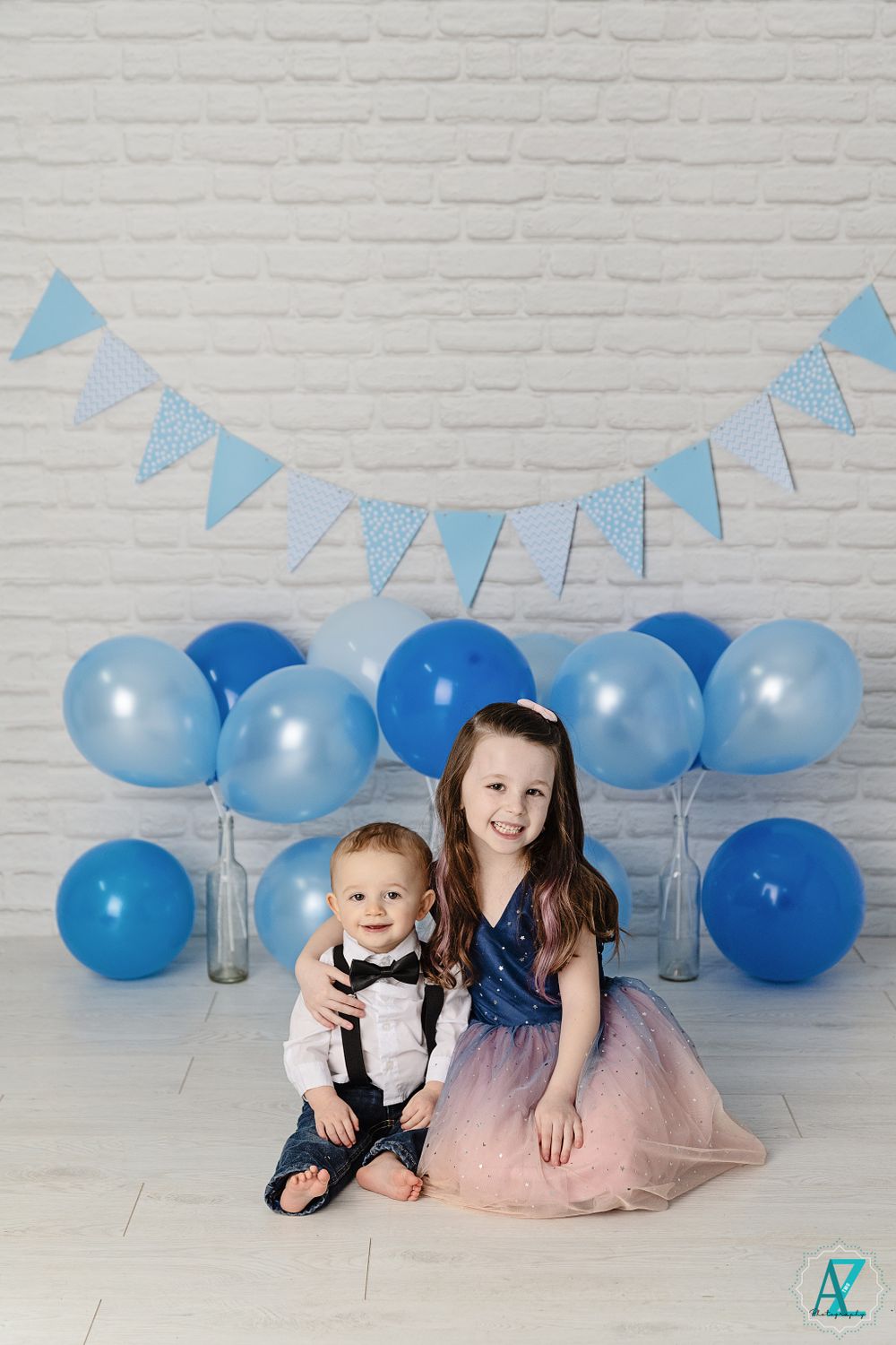 Two children posed with blue balloons and bunting banner against white brick wall.
