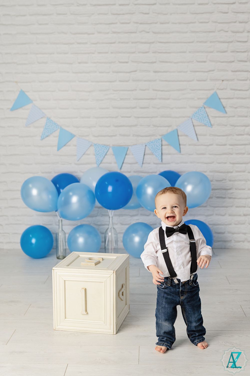 First birthday photo setup with white cube, blue balloons and bunting against brick wall.
