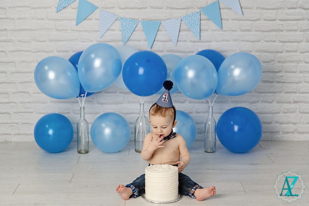Small child with cake and blue balloons for first birthday photoshoot against brick wall.