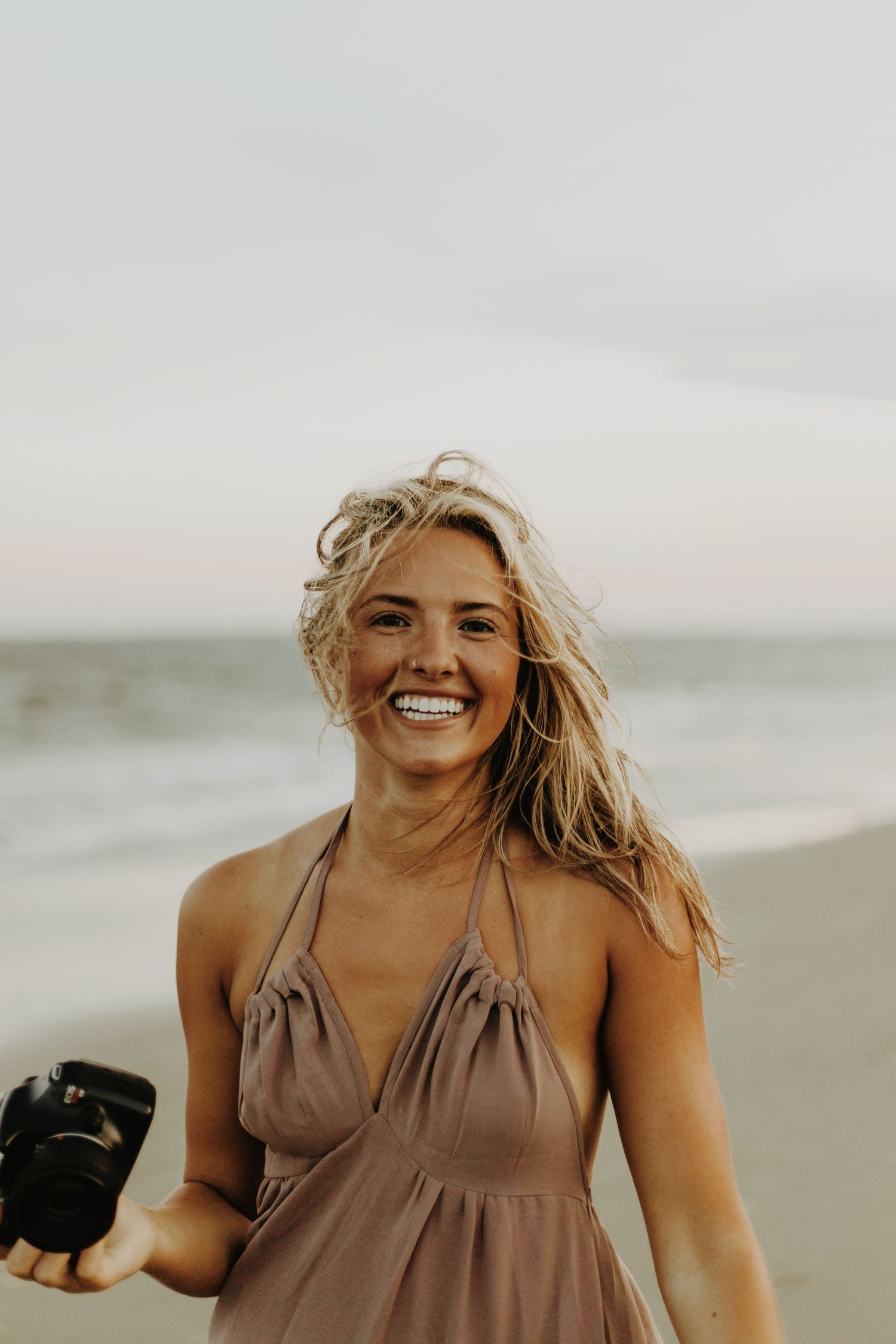 Professional photographer holding a camera smiles joyfully on a cloudy beach in a brown halter dress.