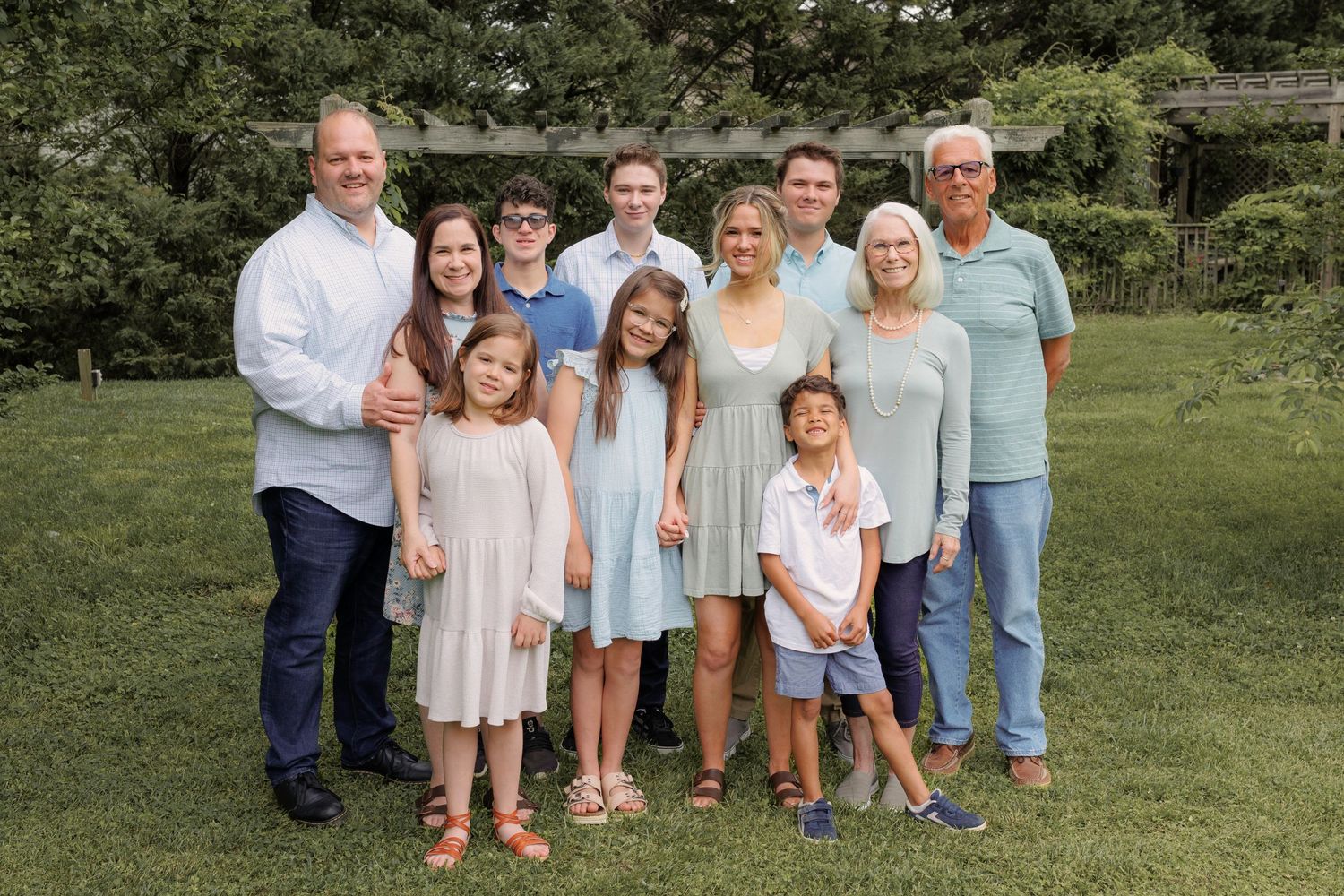 Multi-generational family poses together outdoors in coordinated pastel outfits on a grassy lawn.