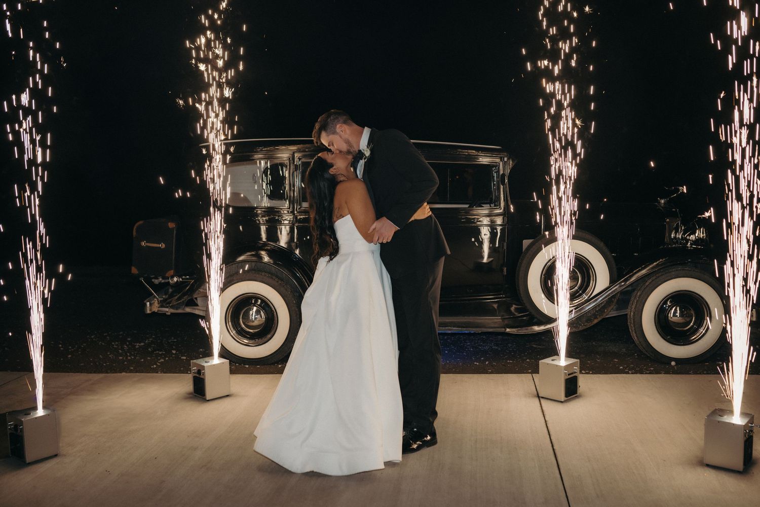 A couple dances surrounded by sparkler fountains with a vintage car in the background.