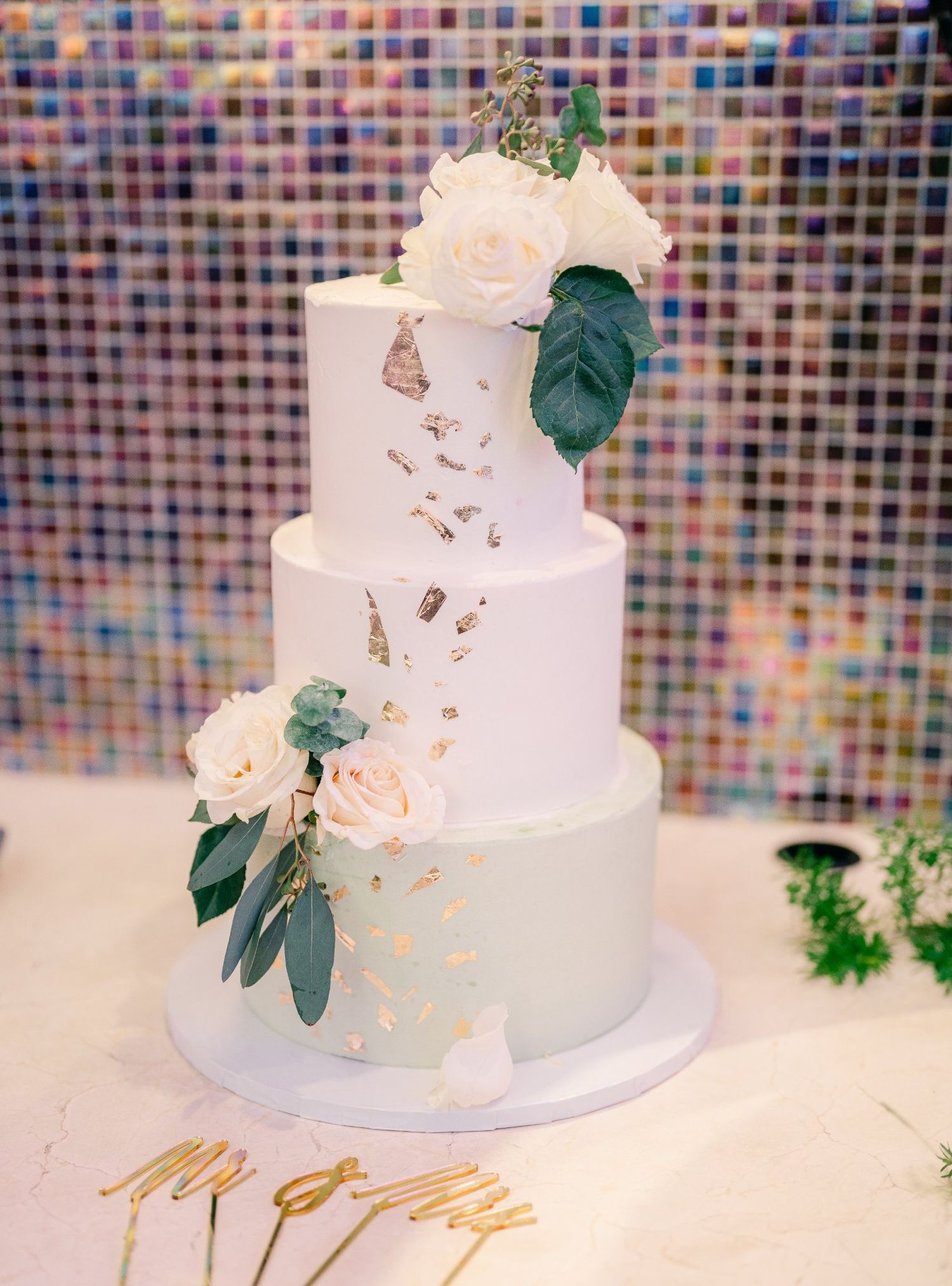 Three-tier white wedding cake decorated with fresh flowers and gold accents against mosaic background.
