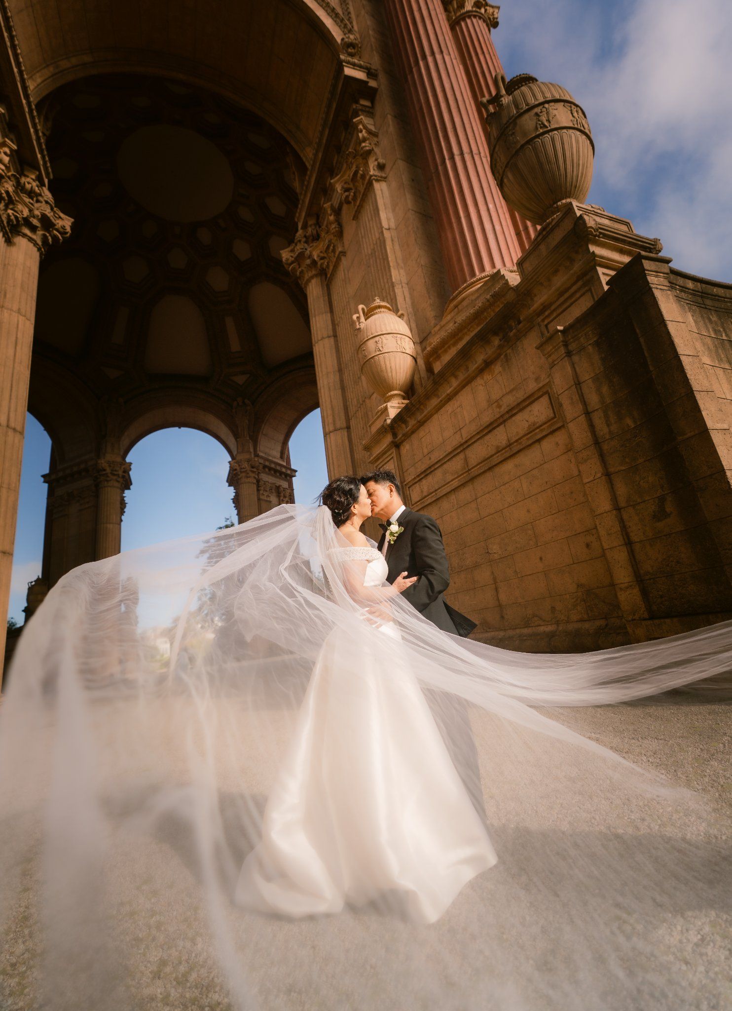Dramatic wedding photo under classical architecture with flowing white veil caught in the wind.