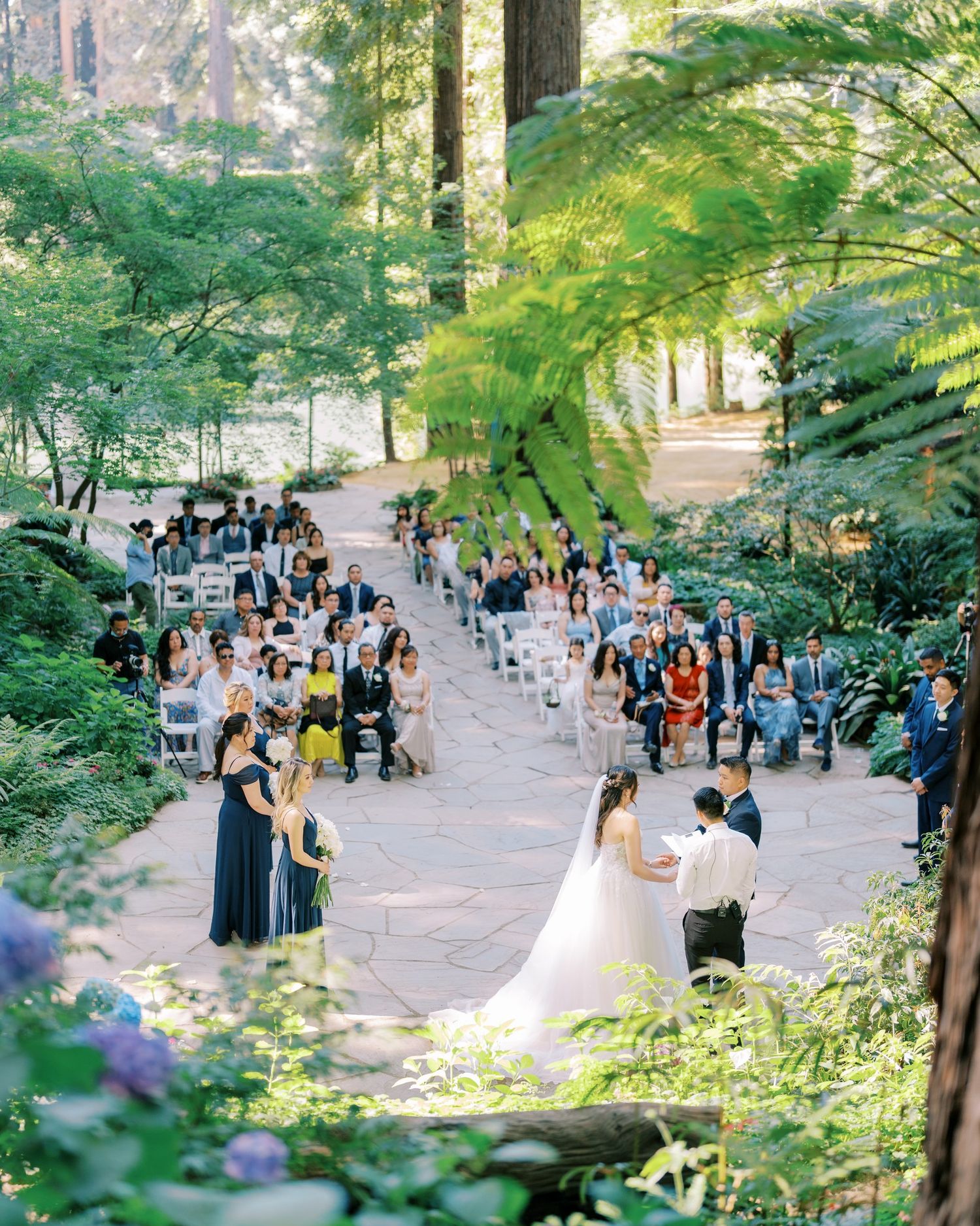 An outdoor forest wedding ceremony with guests seated along a winding path amongst tall trees.