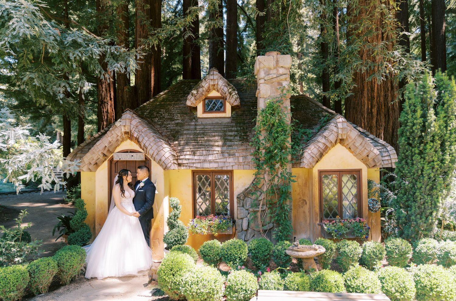 A wedding photo taken in front of whimsical cottage-style buildings surrounded by lush gardens.
